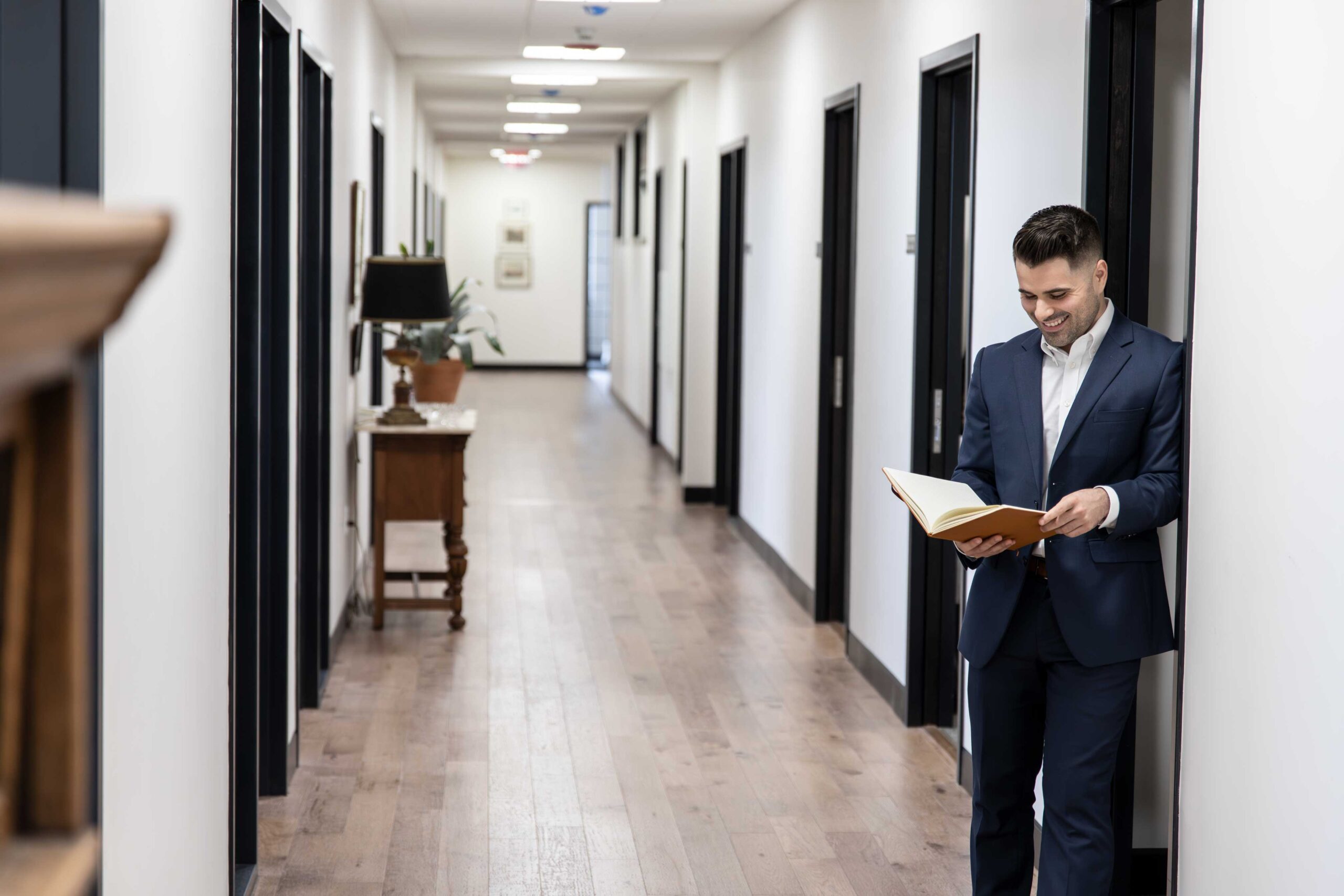 Armando, a man in a navy blue suit, stands in an empty hallway, leaning against a door frame reviewing some notes in a brown leather notebook